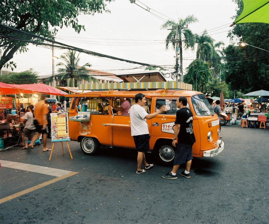 Orange food truck serving refreshments at a bustling Chiang Mai market with vendors and shoppers.