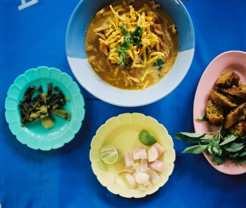 Overhead view of Thai dishes including khao soi noodle soup and sausages in colorful bowls on blue surface.
