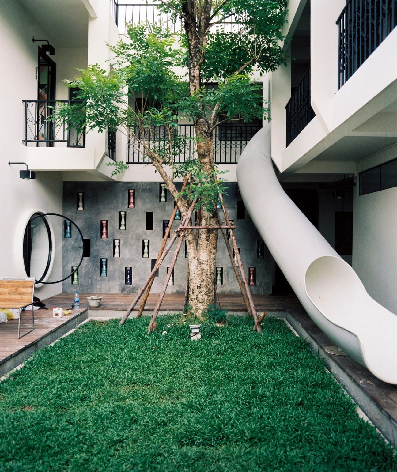 Modern courtyard with multi-story white and grey building featuring green balconies, a large tree, and artificial turf.