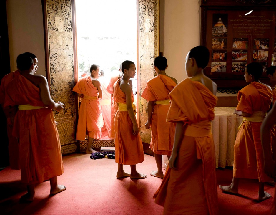 Novice Buddhist monks in orange robes gather inside Wat Phra Singh temple with ornate golden door panels.
