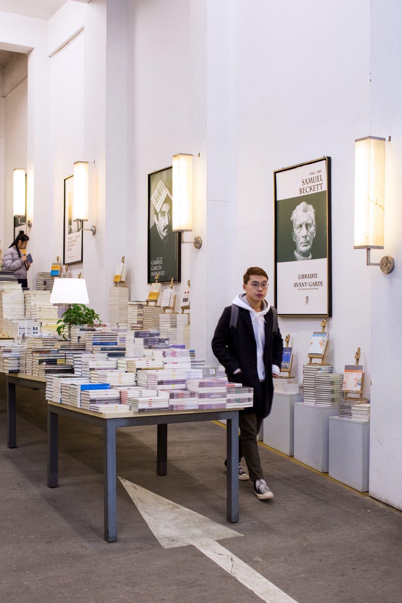 Young man standing in modern bookshop interior with stacked books on display table and wall-mounted author portraits.