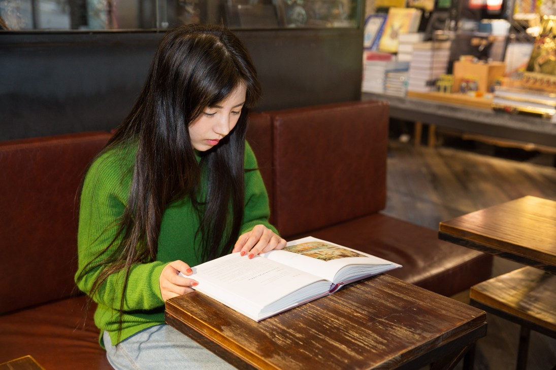 Woman in green sweater reading at table in bookshop café with shelves of books in background.