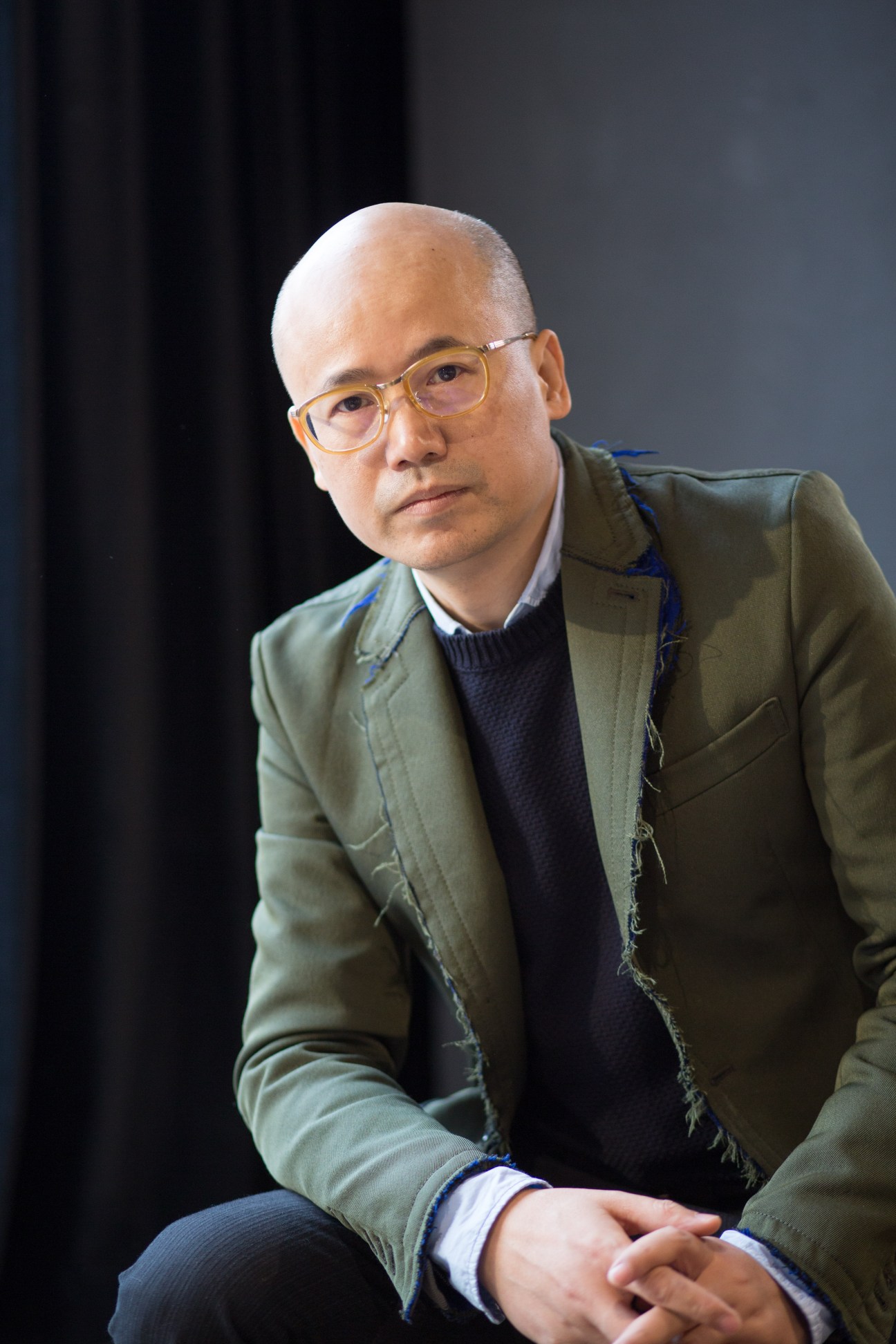 Portrait of Mix Place owner Mao Jihong wearing glasses and an olive jacket against a dark background.