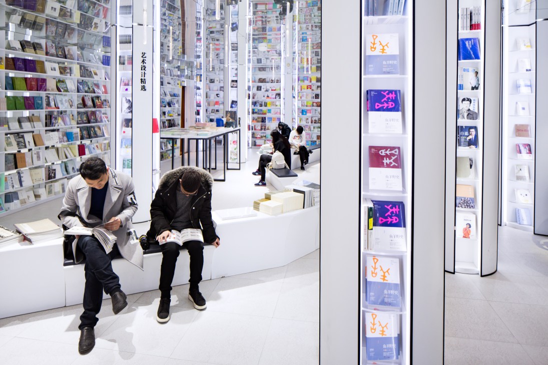 Two customers browse books while seated in Zhongshuge's minimalist white interior with floor-to-ceiling shelving.
