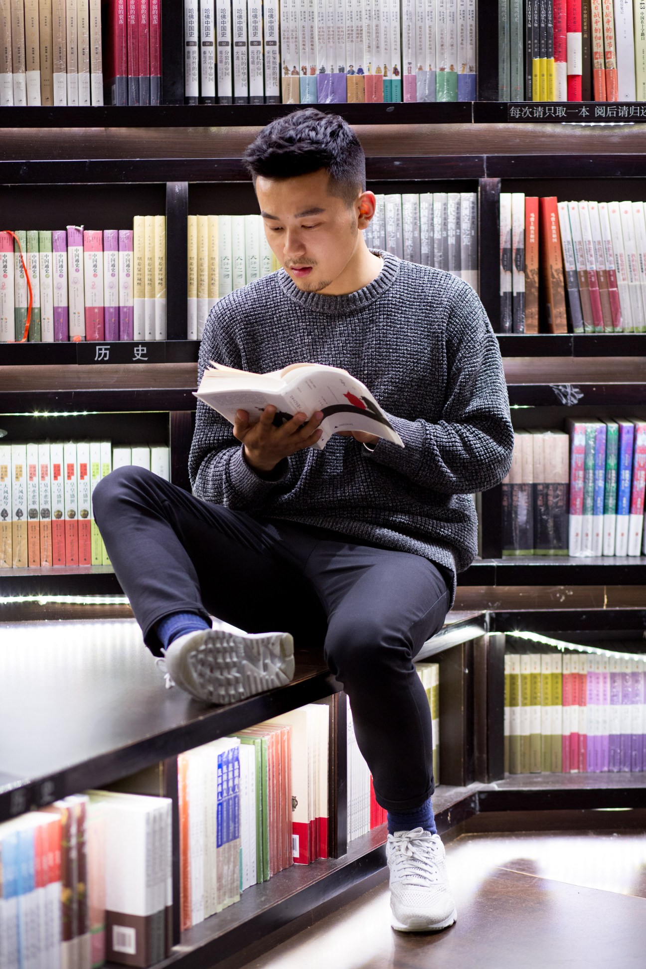 Young man reading a book while seated on a low shelf in a modern Chinese bookshop with colorful volumes behind him.