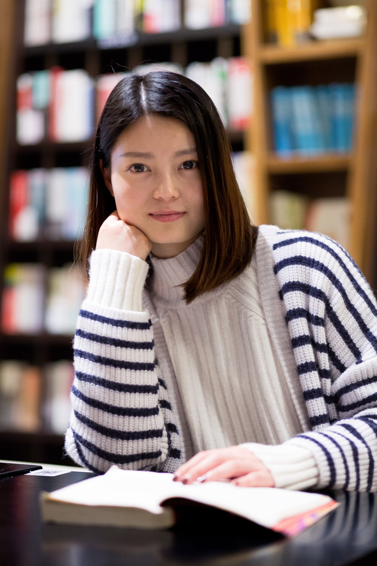 Woman in striped sweater reading at table in bookshop with shelves behind her.