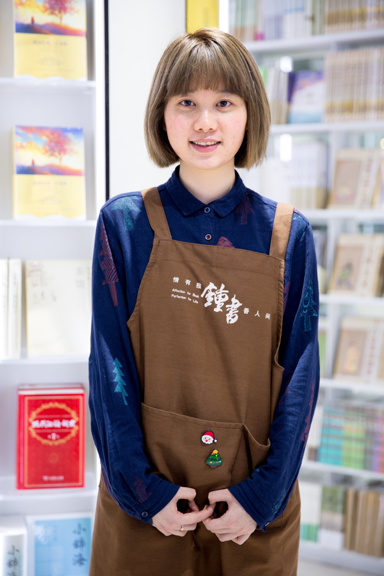 Young female employee wearing brown apron with Chinese characters stands smiling in front of bookshelves at Zhongshuge bookstore.