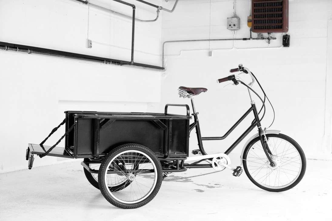 Modern redesigned Sanitov trishaw with stainless-steel frame, leather seat and electric motor in white gallery space.