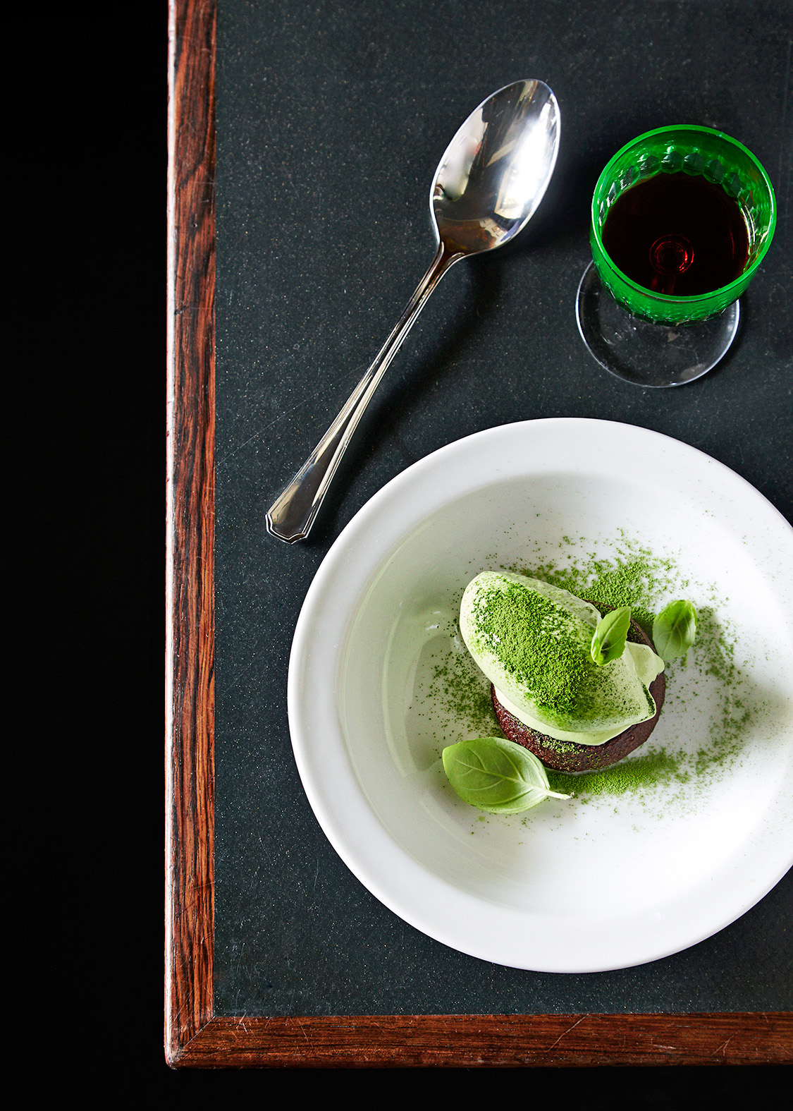A plated dessert with matcha powder, served with a spoon and green drink on dark surface.