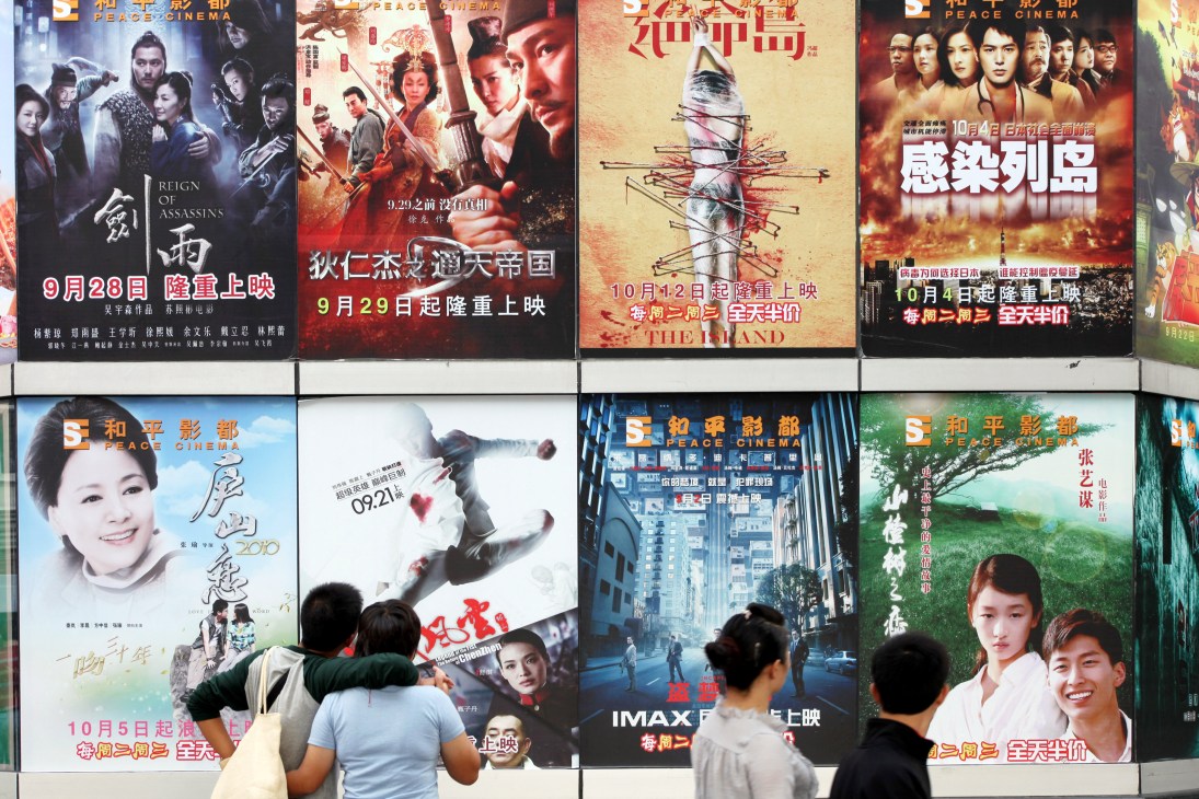 Movie posters with Chinese characters displayed on a cinema wall with people viewing them.