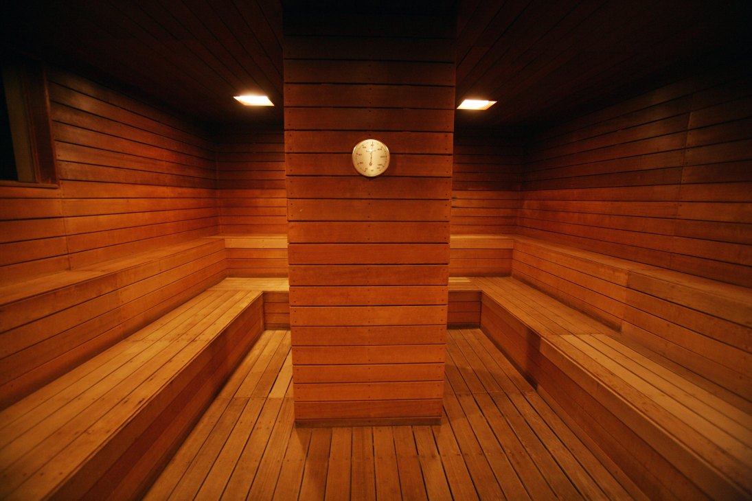 Interior of a wooden sauna with bench seating, a central wooden pillar, wall-mounted clock, and recessed lighting.