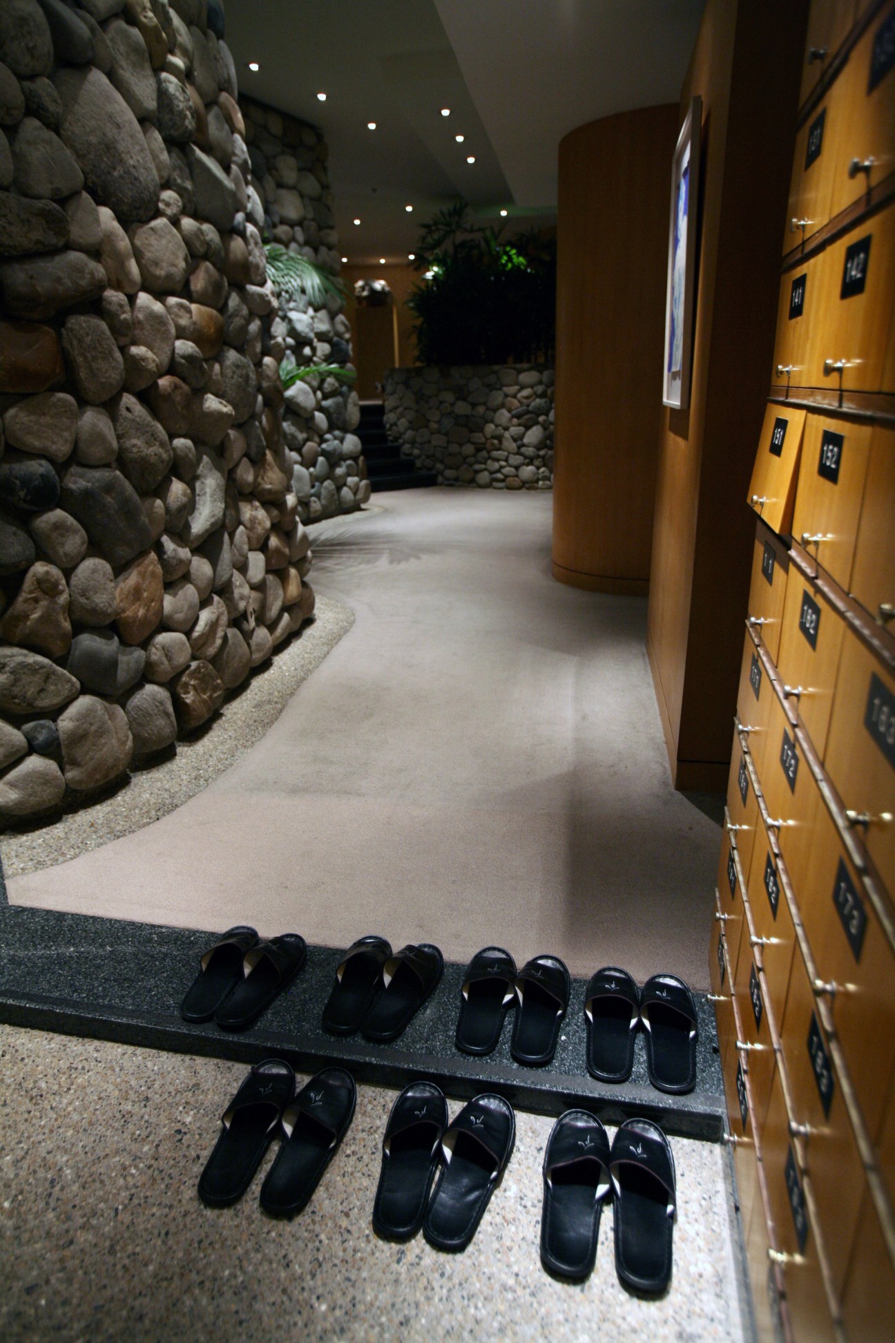 Interior corridor of spa locker room with stone walls, wooden lockers, and black slippers arranged on floor.