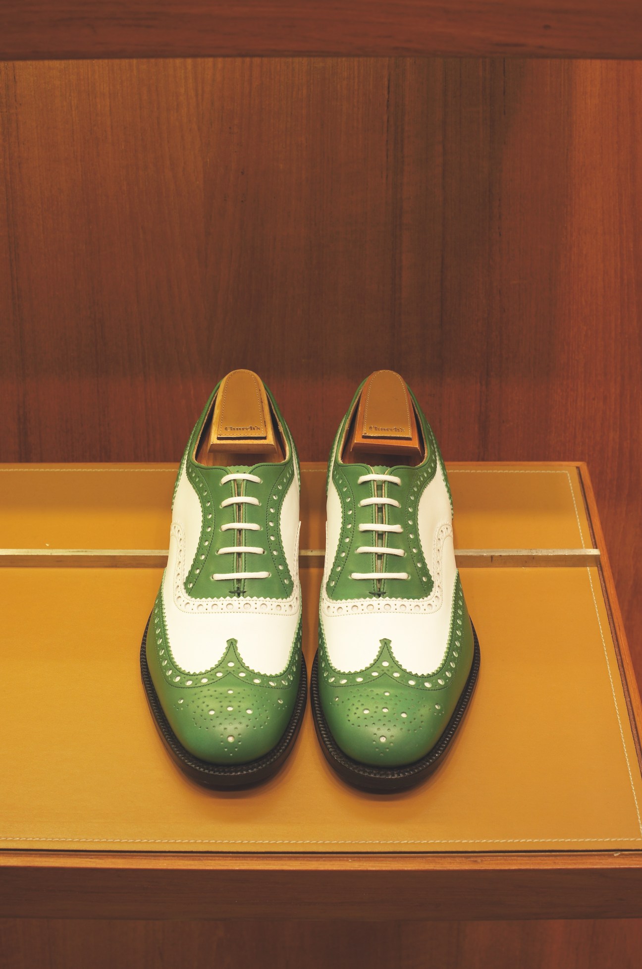 Pair of green and white leather brogue oxford shoes by Church's displayed on a yellow leather desk pad.