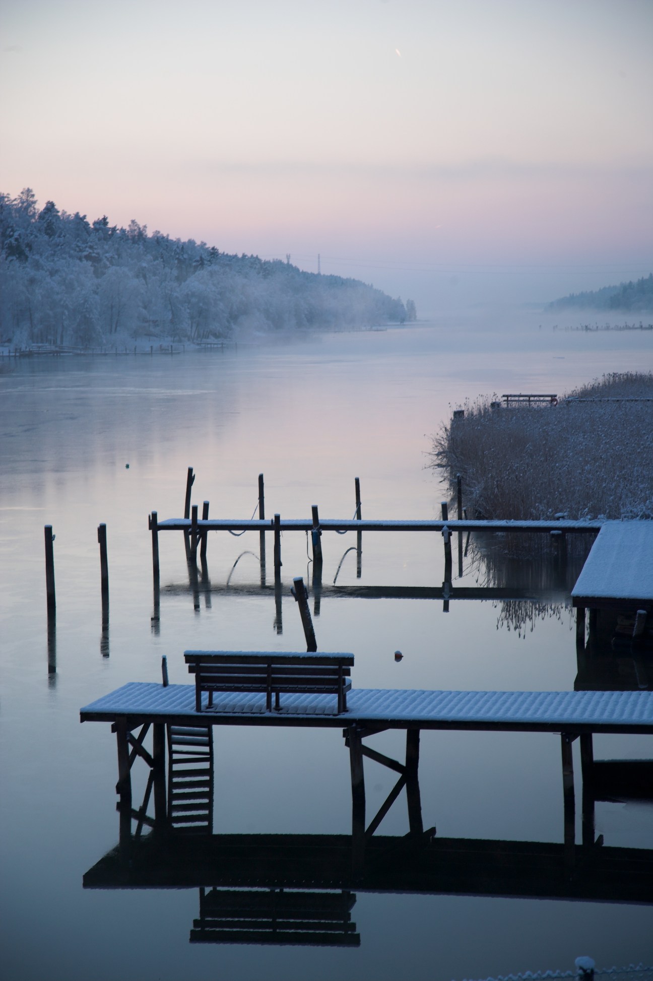 Frost-covered wooden docks and benches on a frozen Finnish lake at dawn, with snow-laden forest and mist in the background.