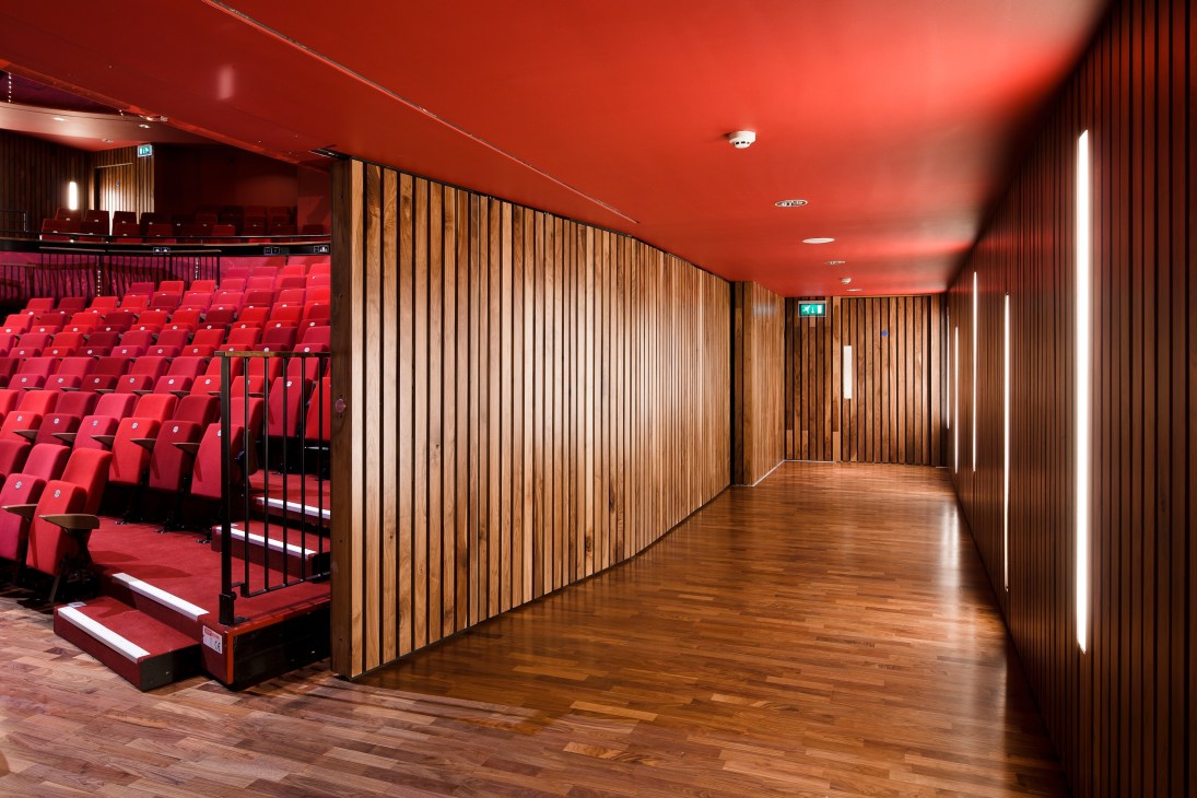 Modern corridor with red ceiling, wooden slat walls, and theater seating visible through glass partition.