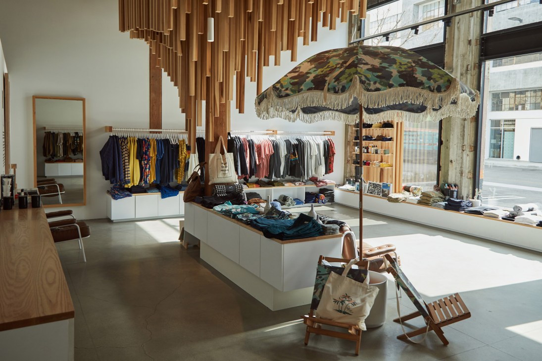 Interior of Banks Journal menswear shop with clothing racks, white display cases, wooden ceiling feature, and patterned umbrella with fringe.