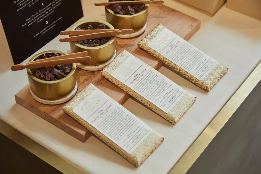 Gold bowls filled with chocolate pieces and wooden chopsticks, alongside packaged chocolates with patterned wrappers on a wooden surface.