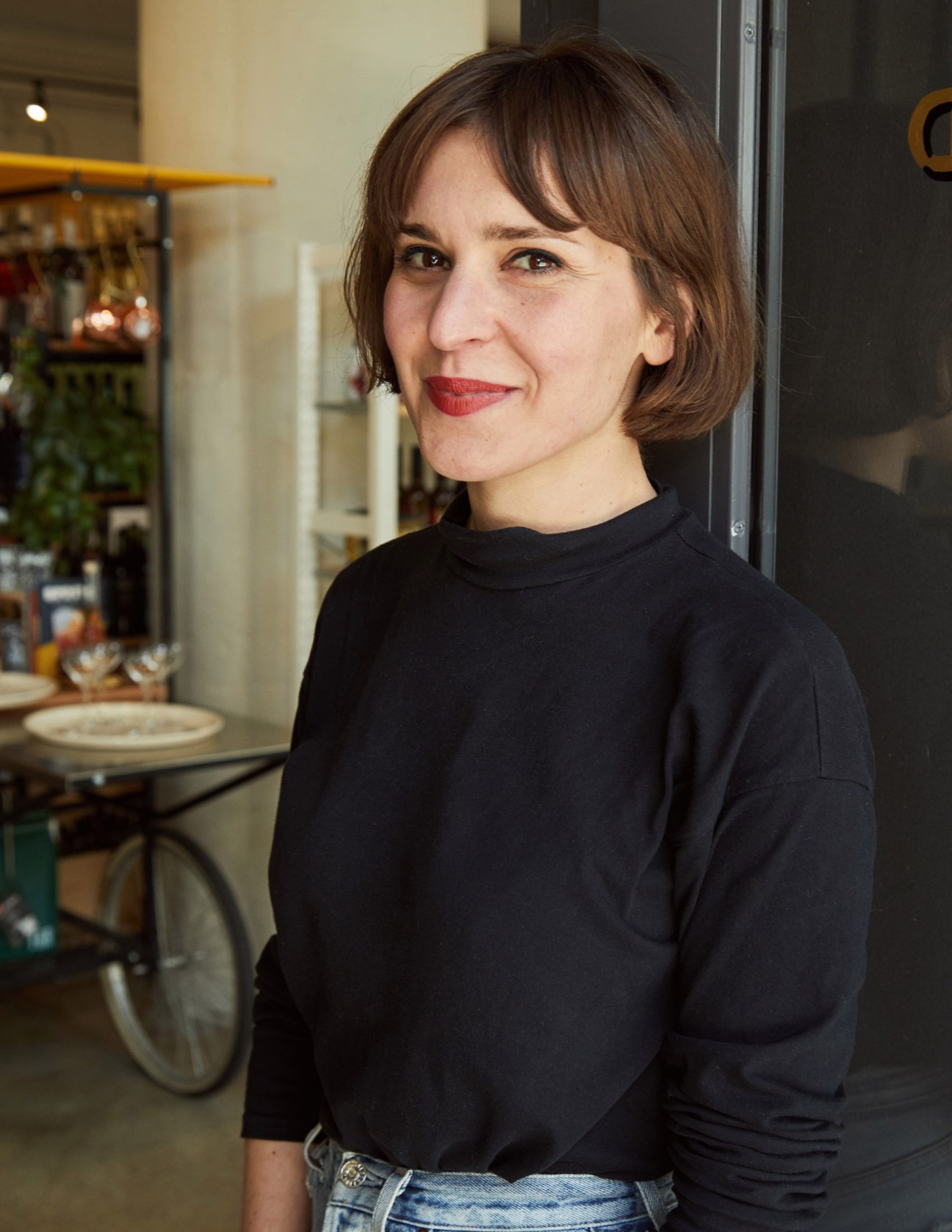 Woman with brown hair and red lipstick smiling at camera inside Flask & Field store with shelves and bar visible in background.
