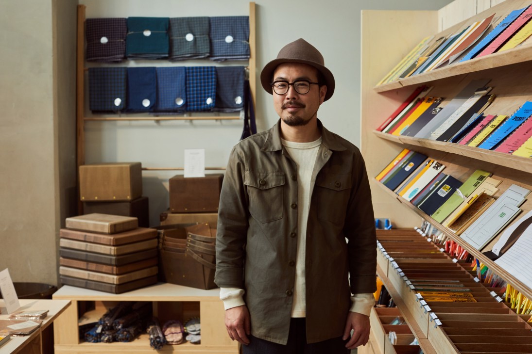 Yuichi Munehiro, founder of Hightide Store, standing in his stationery shop surrounded by organized shelves of notebooks and supplies.