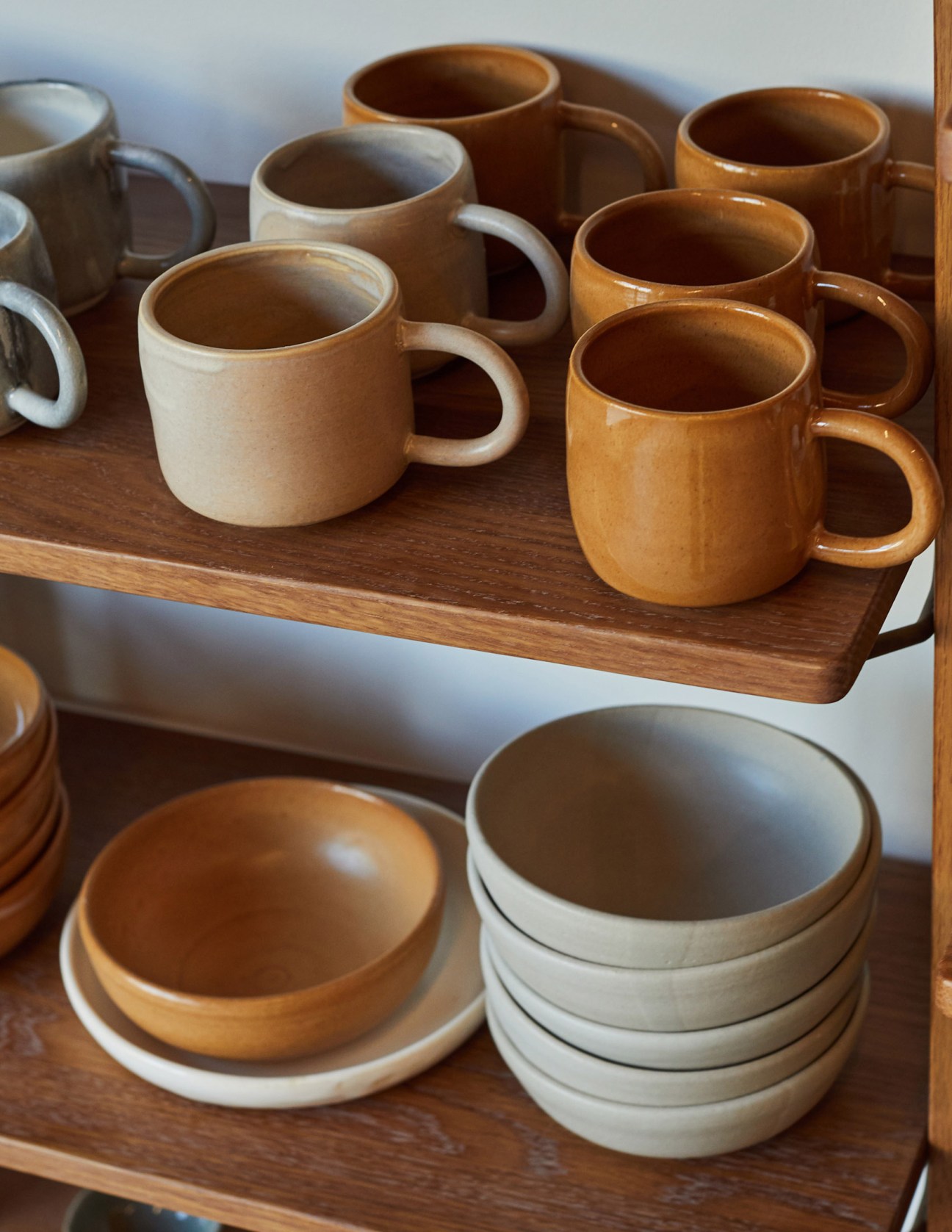 Hand-thrown ceramic mugs in cream and brown tones displayed on wooden shelves.