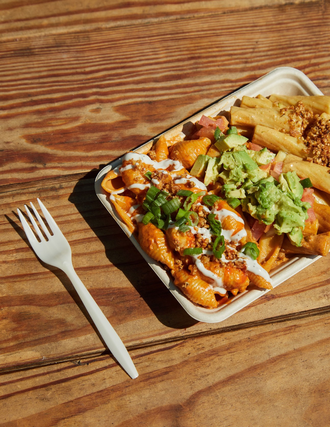 A white paper container with loaded fries topped with guacamole, crumbled meat, and green onions on a wooden table with a plastic fork.