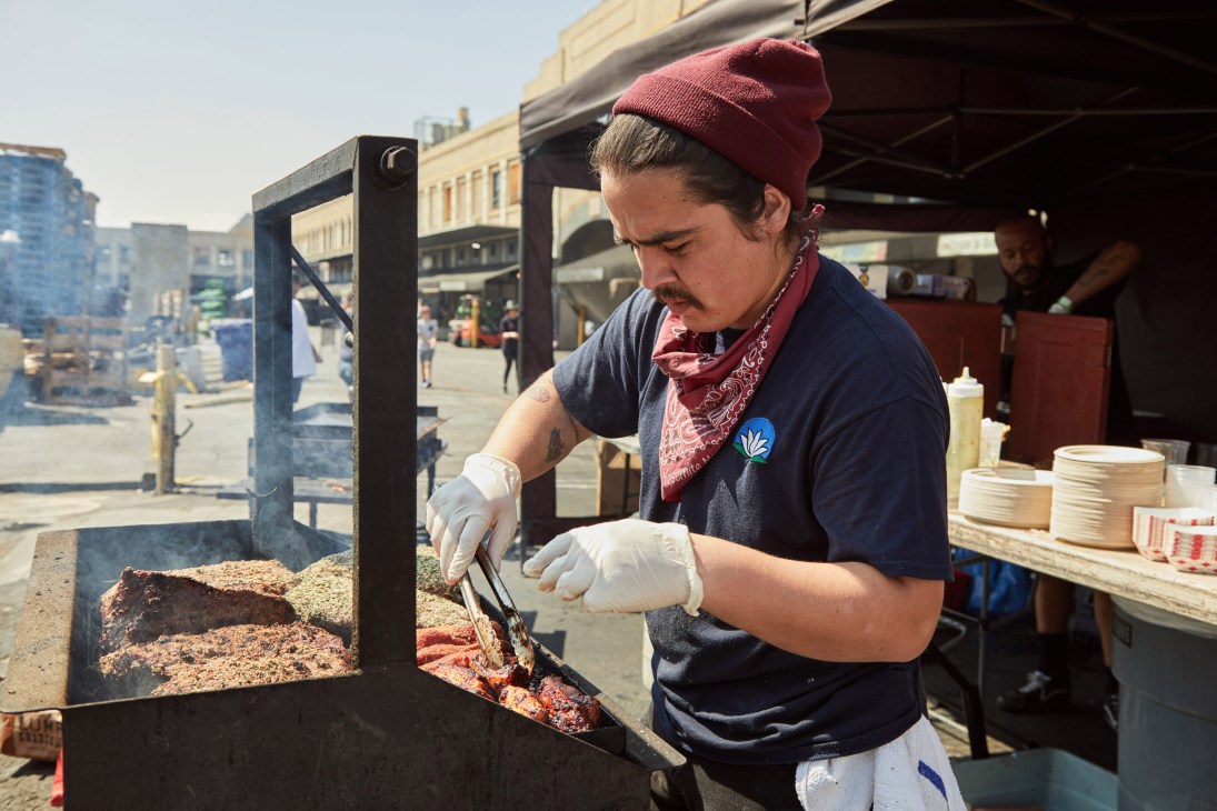 A man in a burgundy beanie and navy shirt prepares meat on a grill at an outdoor food stall.