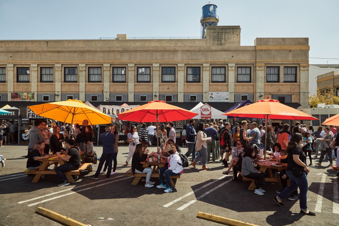 Smorgasburg LA's Sunday market in Downtown Los Angeles with crowds gathered under colorful umbrellas at wooden picnic tables.