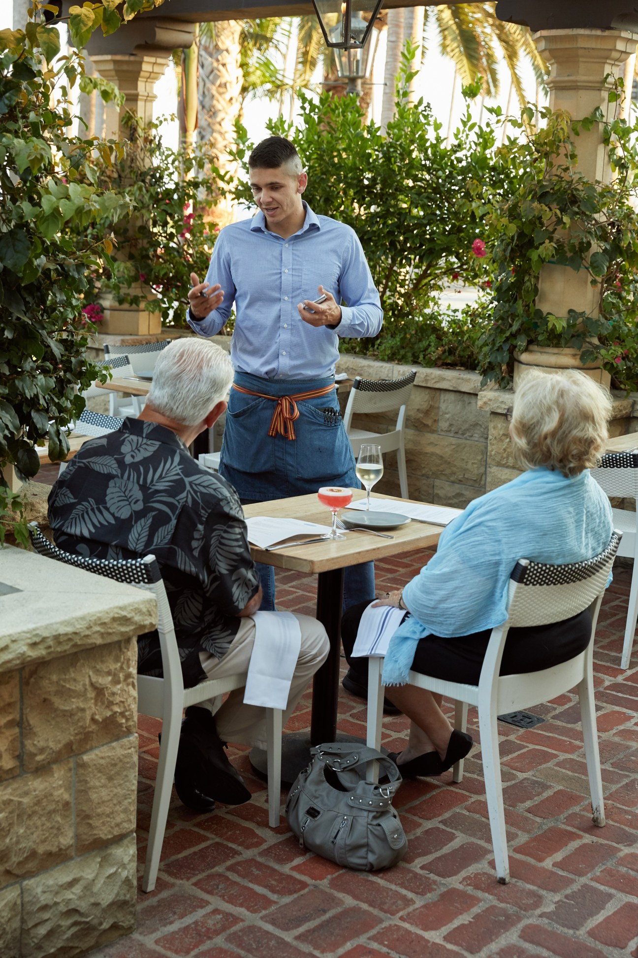 A server in an apron explains menu items to two diners seated at an outdoor restaurant table with wine glasses
