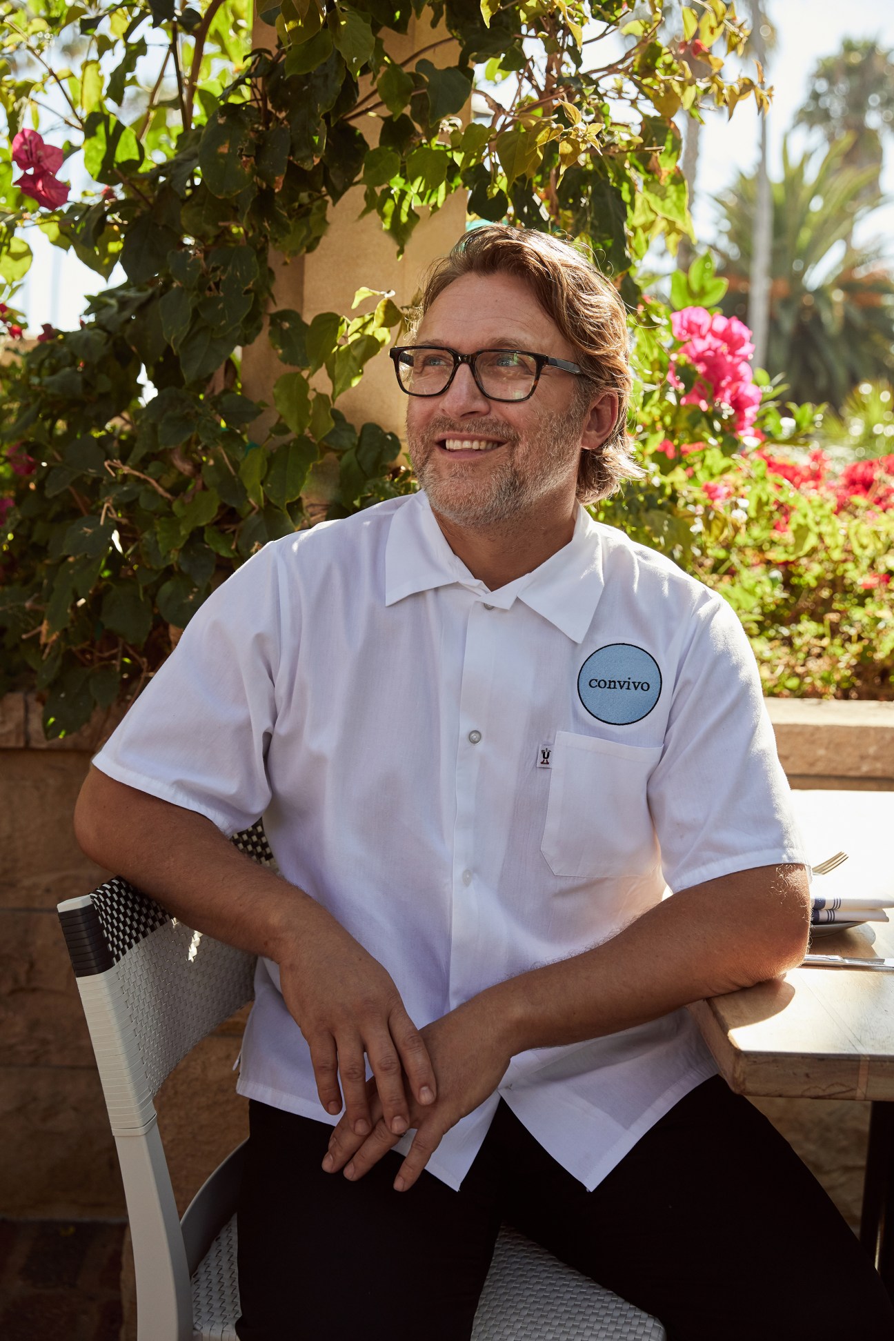 Chef Peter McNee smiling and sitting outdoors wearing glasses and a white Convivo polo shirt