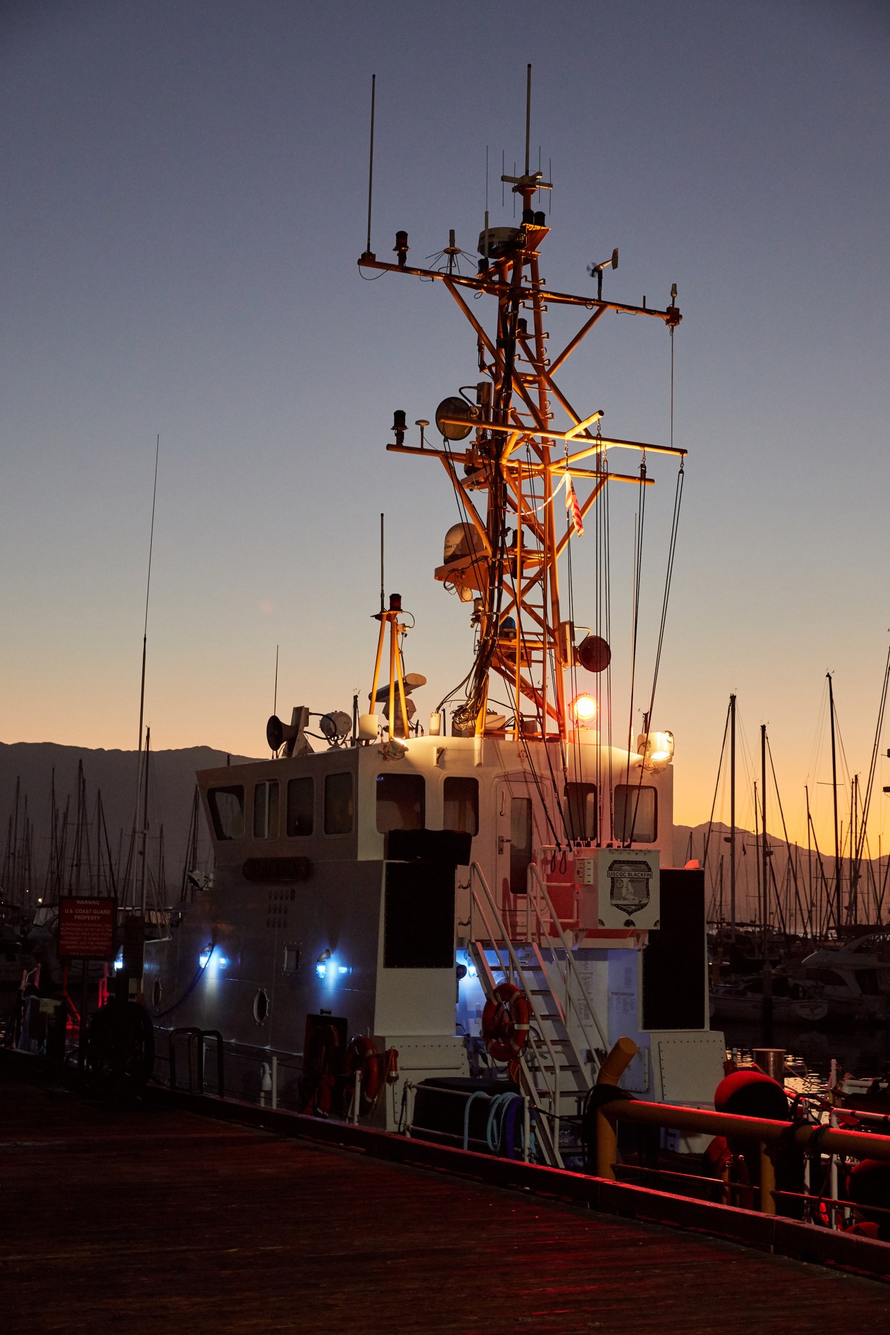 A fishing boat docked in a harbor at sunset, with its mast and lights illuminated against an orange sky.
