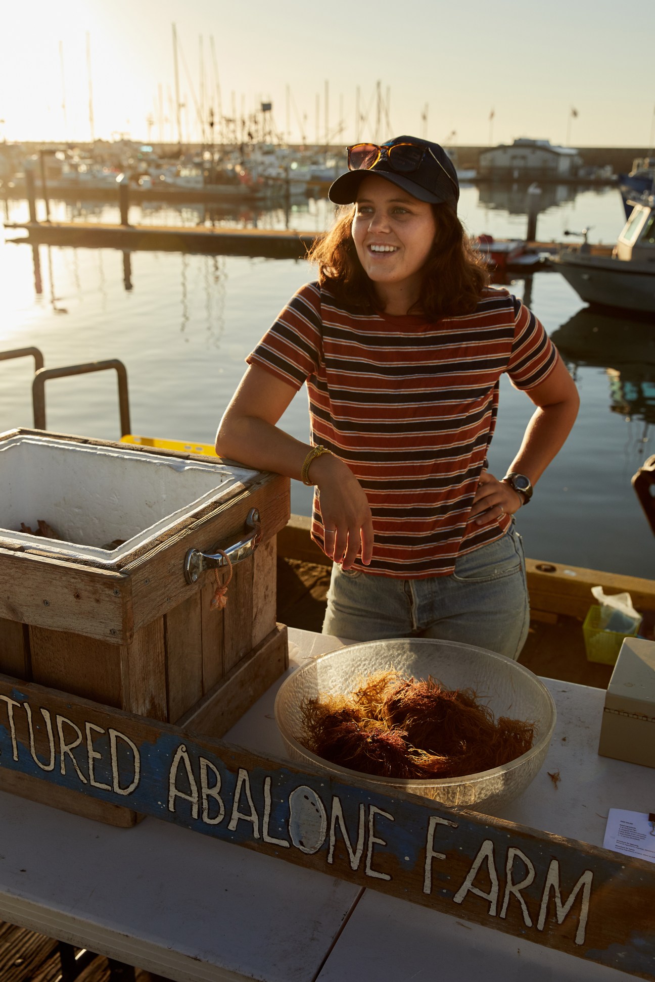A woman in a striped shirt standing by a wooden cooler and bowl of seafood at a sunny dock.