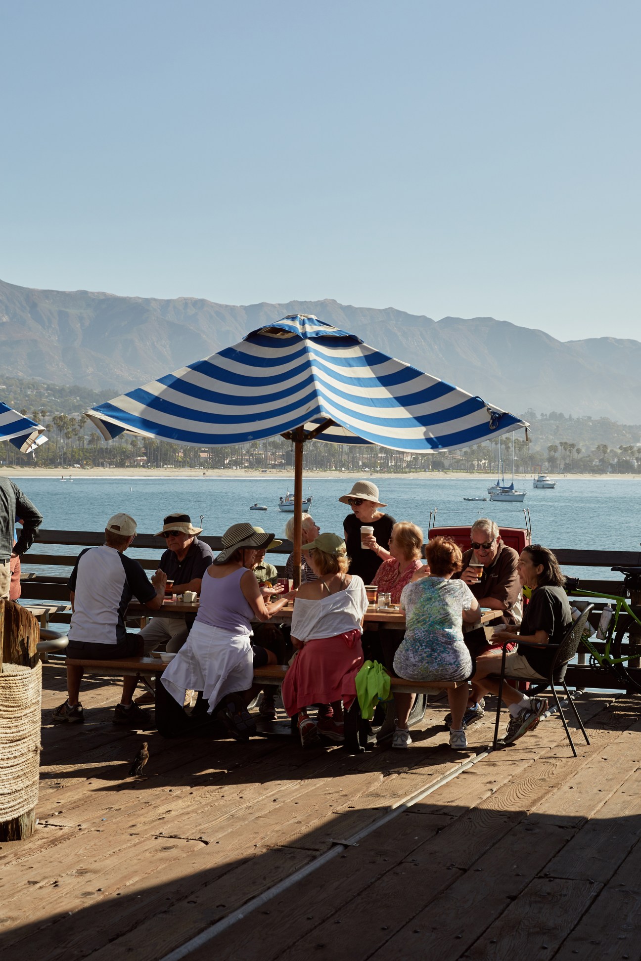 People dining together under a striped umbrella at an outdoor waterfront restaurant with ocean and mountain views