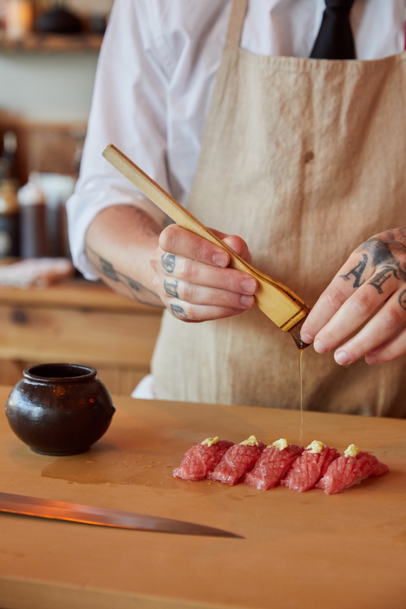 Tattooed hands drizzling dark sauce from wooden utensil onto sliced raw fish at Sushi Bar Montecito