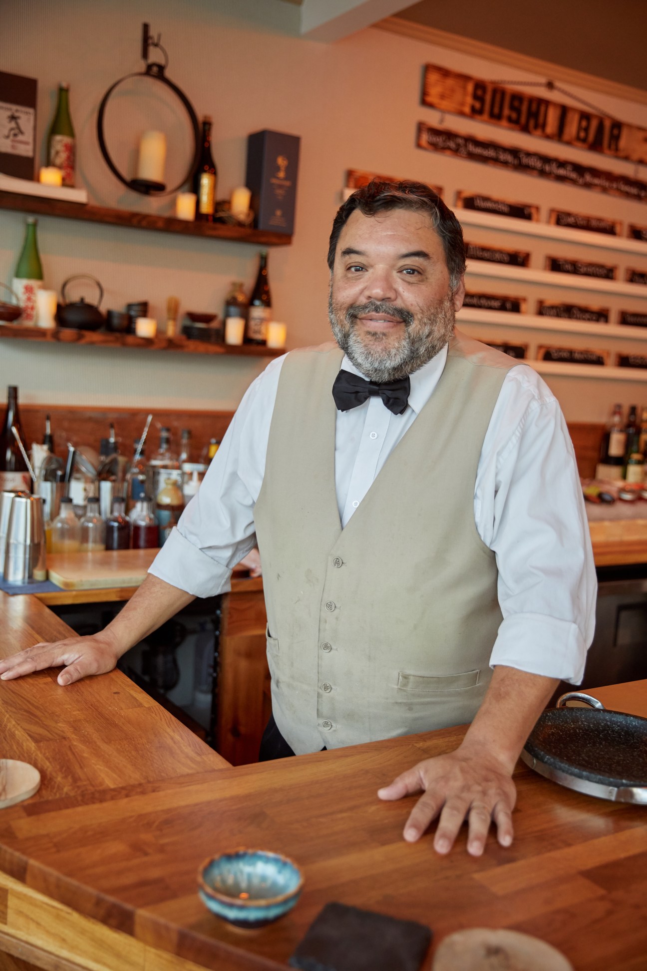 A smiling middle-aged man with a graying beard, wearing a beige vest, white shirt, and black bow tie, standing behind a wooden bar counter.