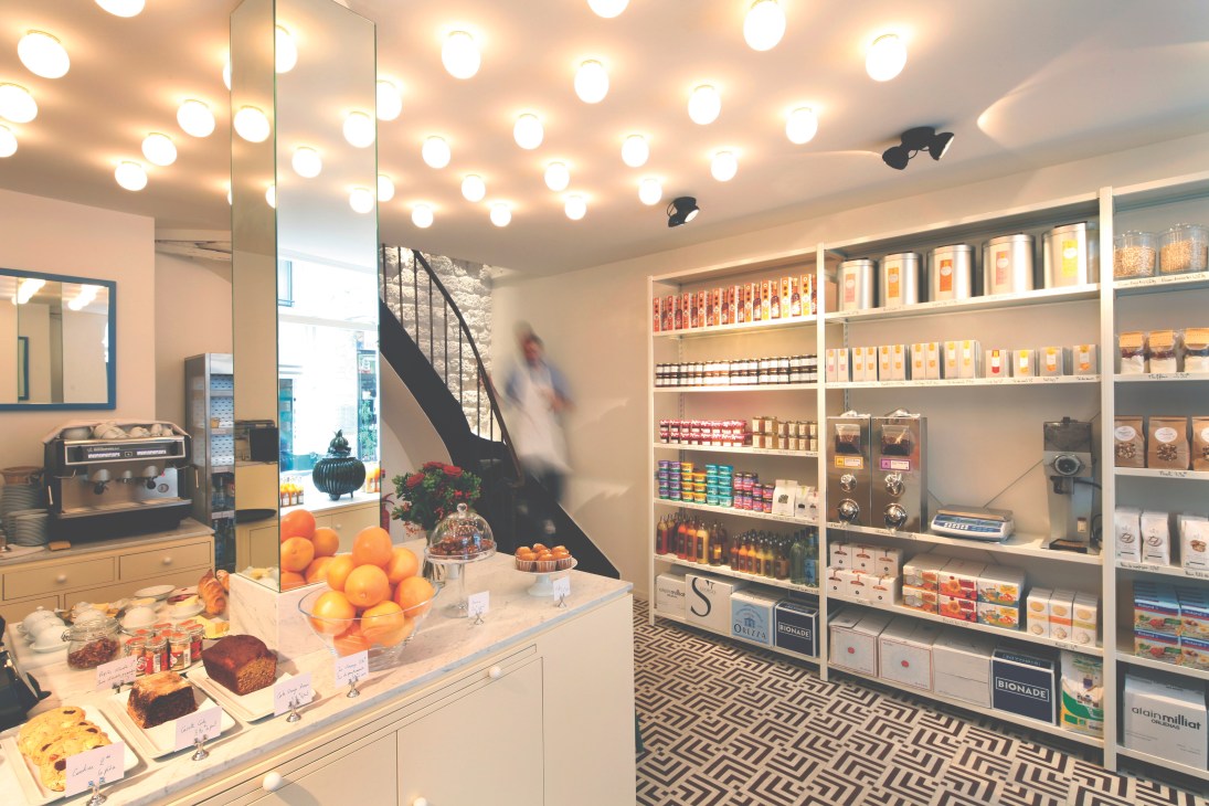 Interior of Claus deli in Paris with a counter display of fresh pastries, oranges, and breakfast items, shelving stocked with ingredients and products, and modern lighting fixtures overhead.