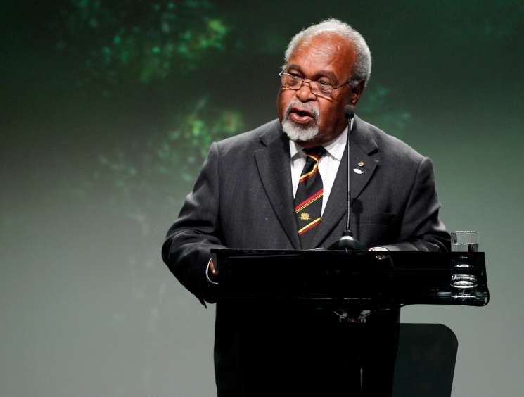 Sir Michael Somare, Papua New Guinea's founding father, speaking at a podium wearing glasses and a dark suit with a colorful tie.