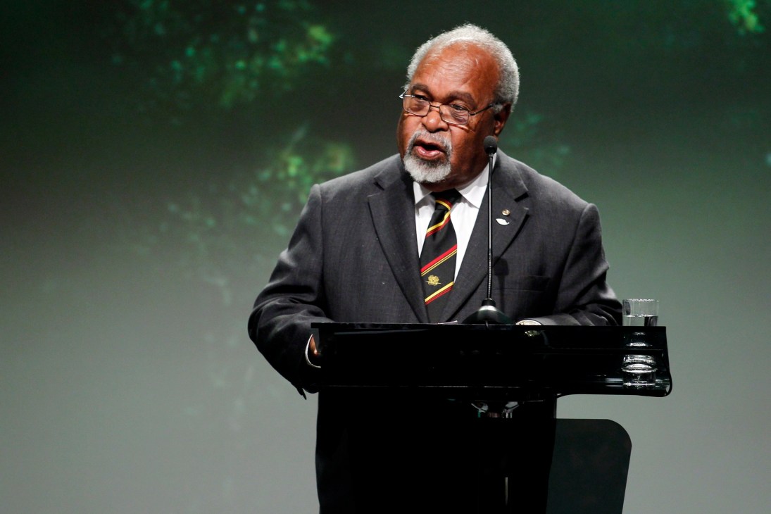 Sir Michael Somare, Papua New Guinea's founding father, speaking at a podium wearing glasses and a dark suit with a colorful tie.