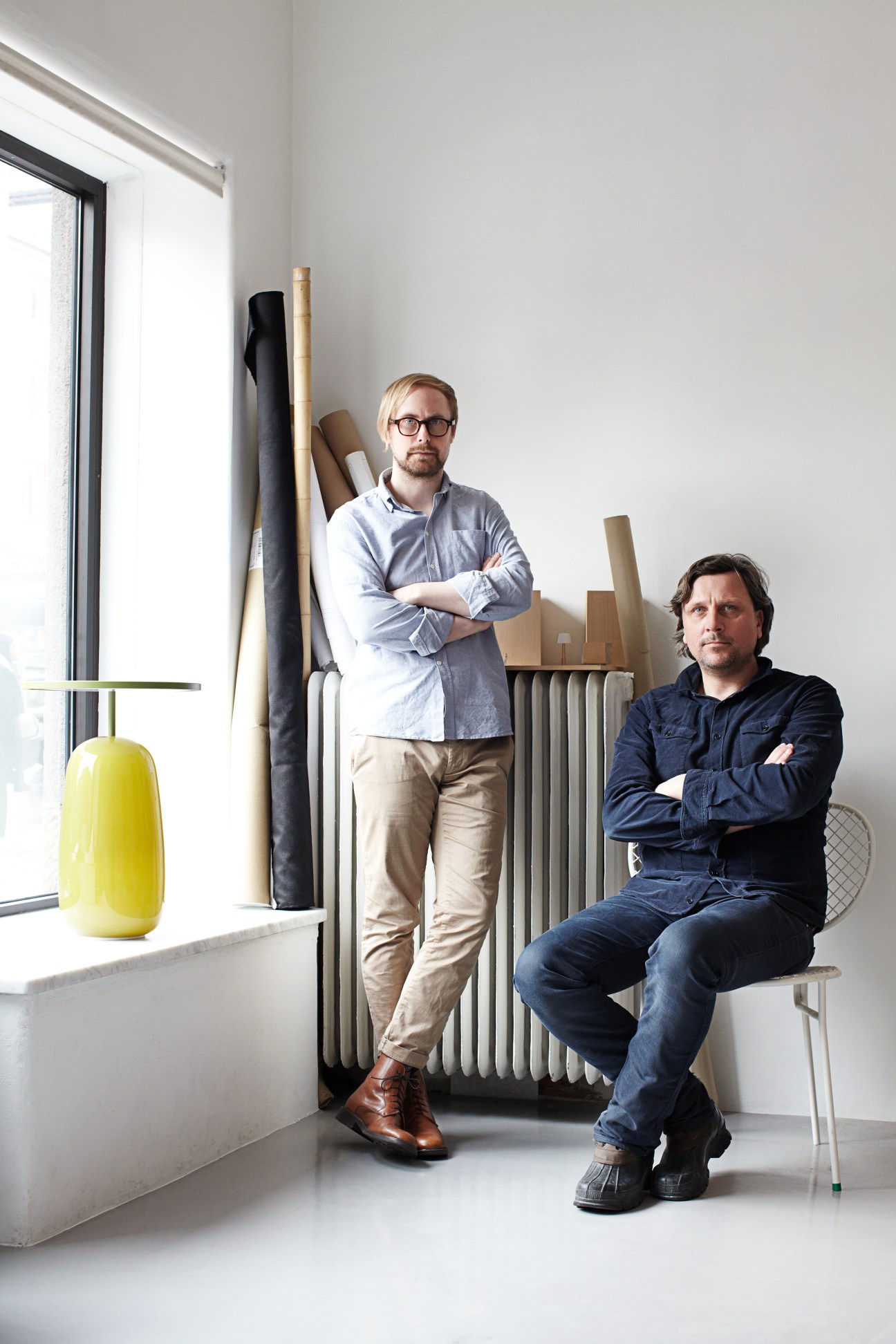 Two male designers posing in a studio with design materials and samples displayed behind them.