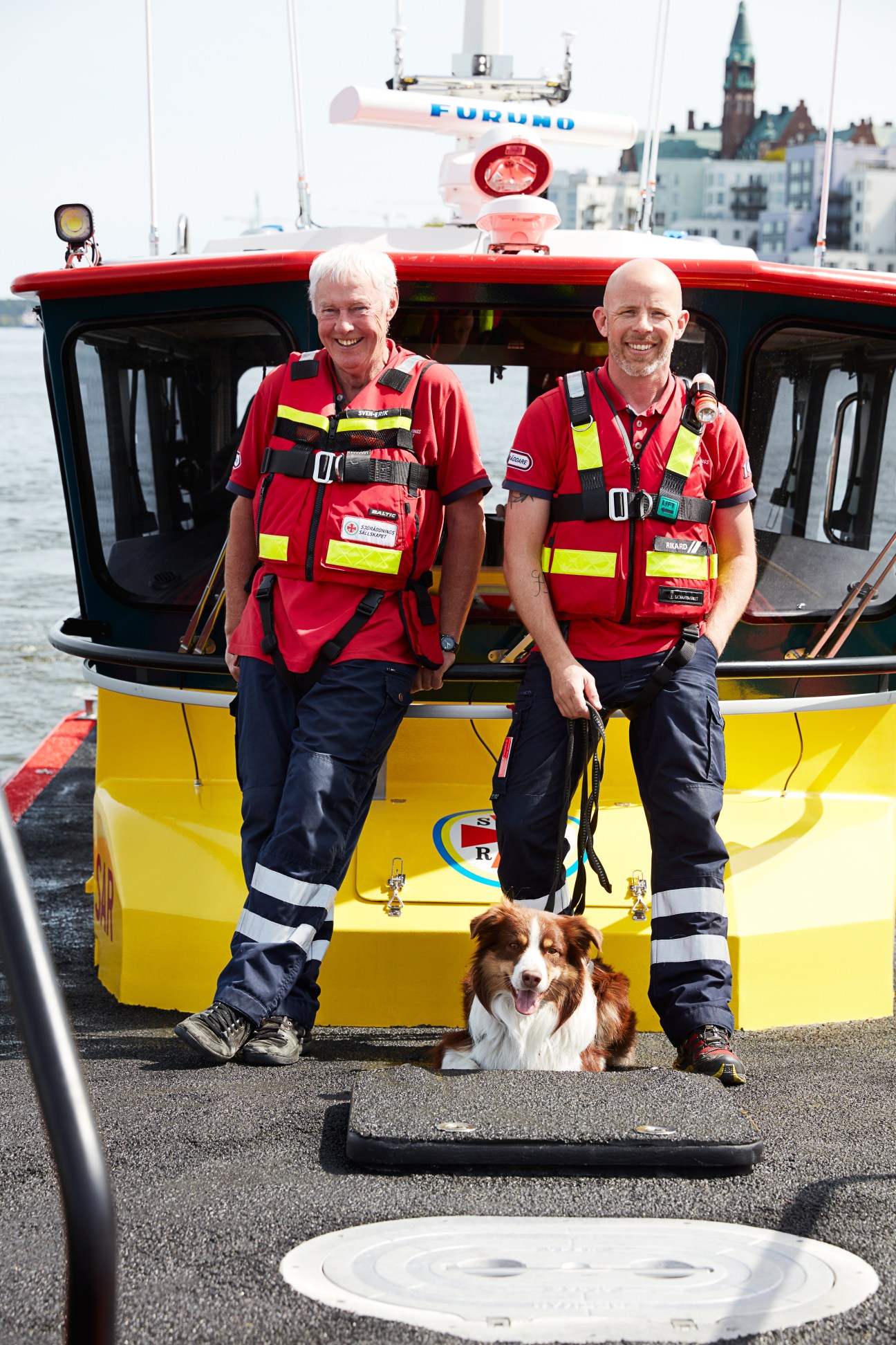 Two Swedish Sea Rescue Society coastguards in red uniforms standing with a rescue dog in front of a yellow and red rescue boat in Stockholm harbor.