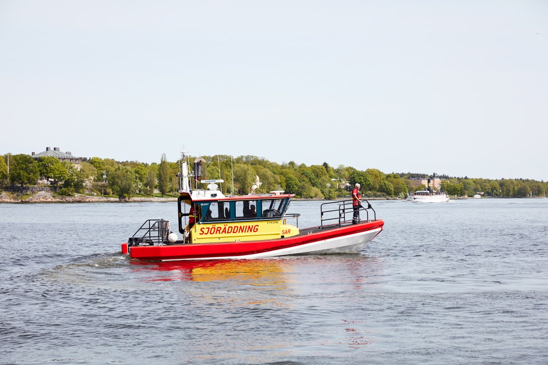 Red and yellow Swedish Sea Rescue Society boat departing from Stockholm waterfront with crew and rescue dog on board.