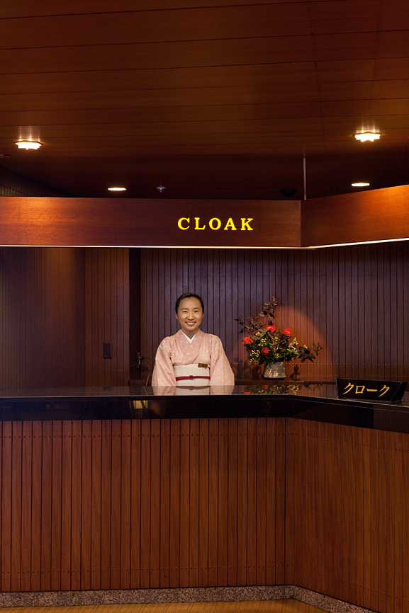 Cloakroom attendant in traditional pink kimono at the Hotel Okura's reception desk with wooden paneling and floral arrangement.