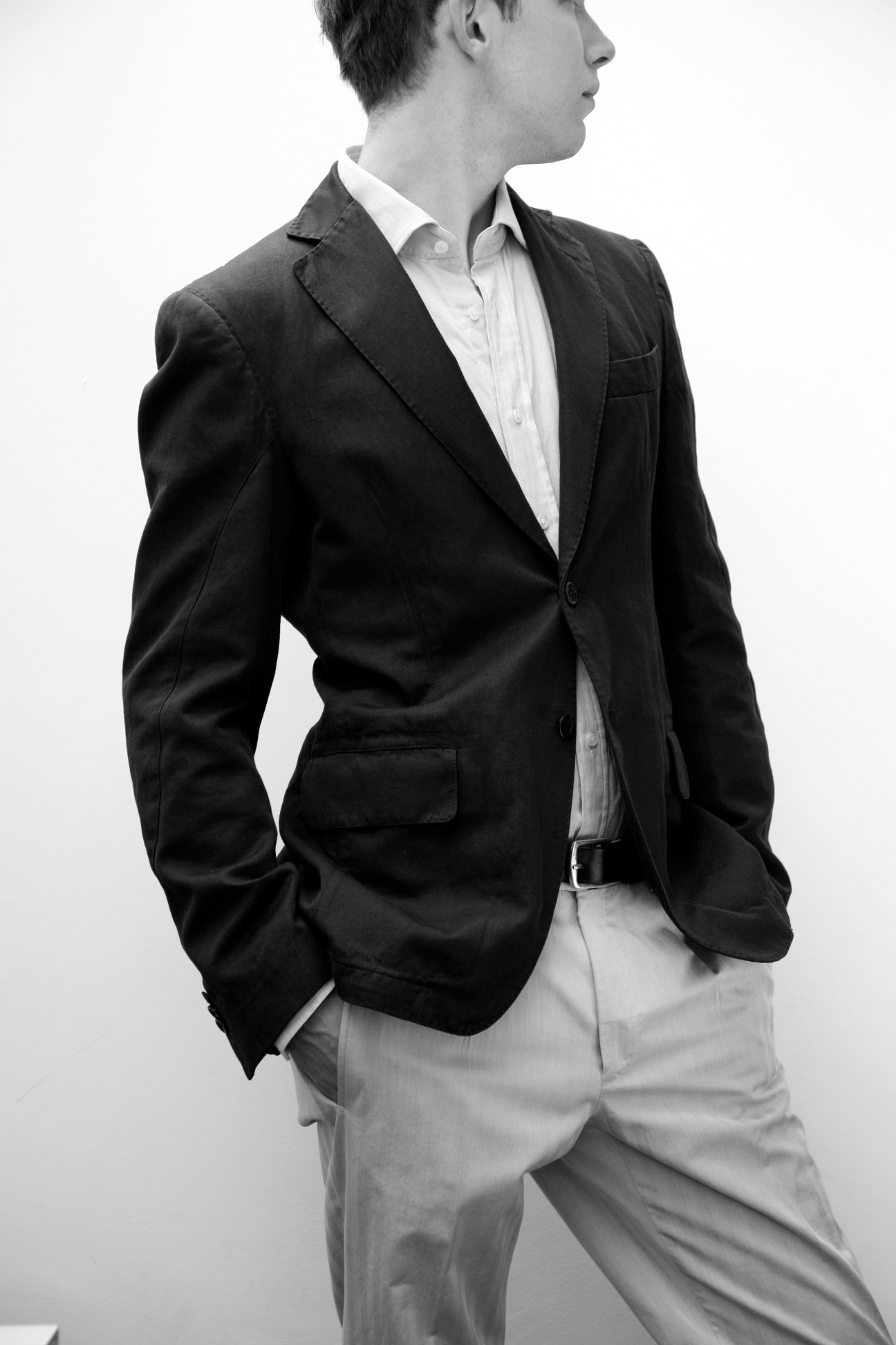 Man wearing black blazer, white shirt, and light gray trousers in profile against white background.