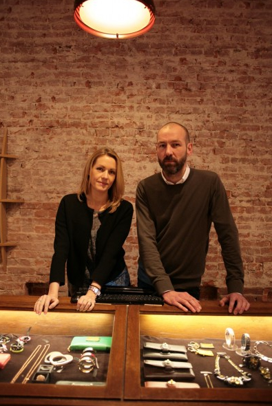 Two people stand behind a jewelry display counter in a brick-walled retail space.