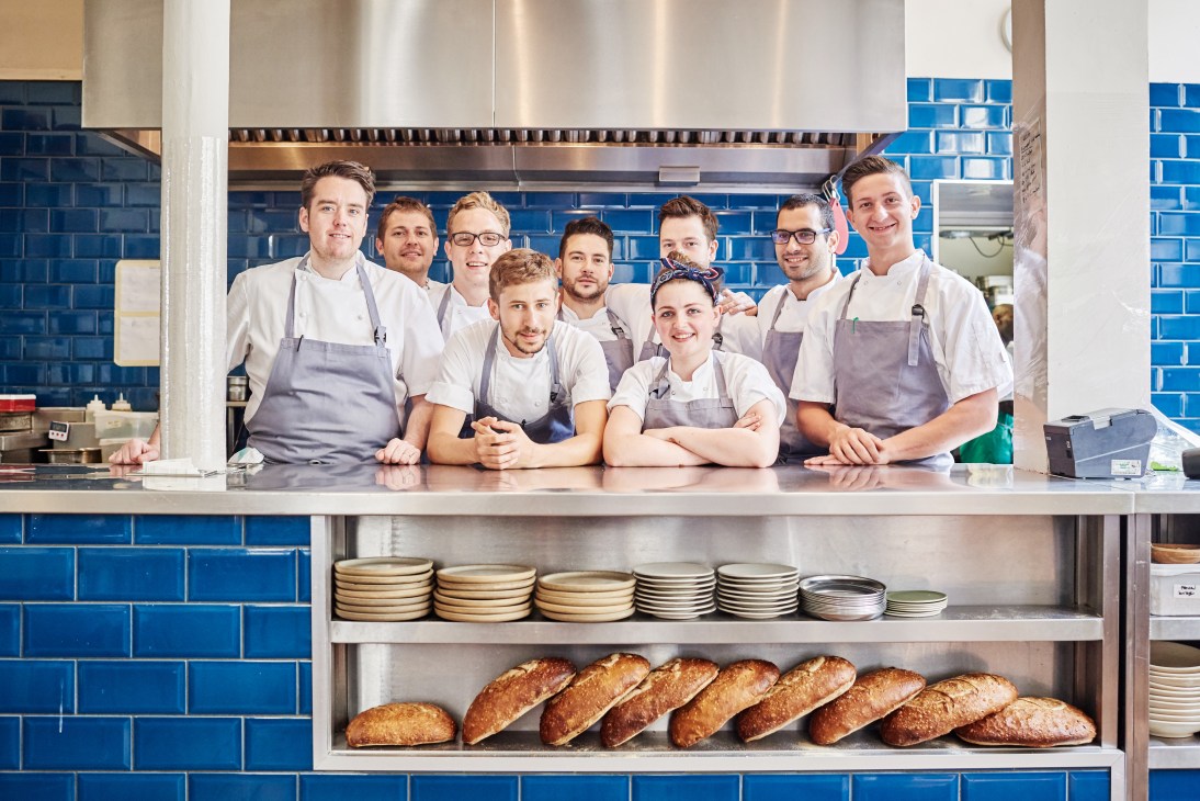 The Clove Club kitchen staff in uniform stand behind counter with bread and plates displayed below.