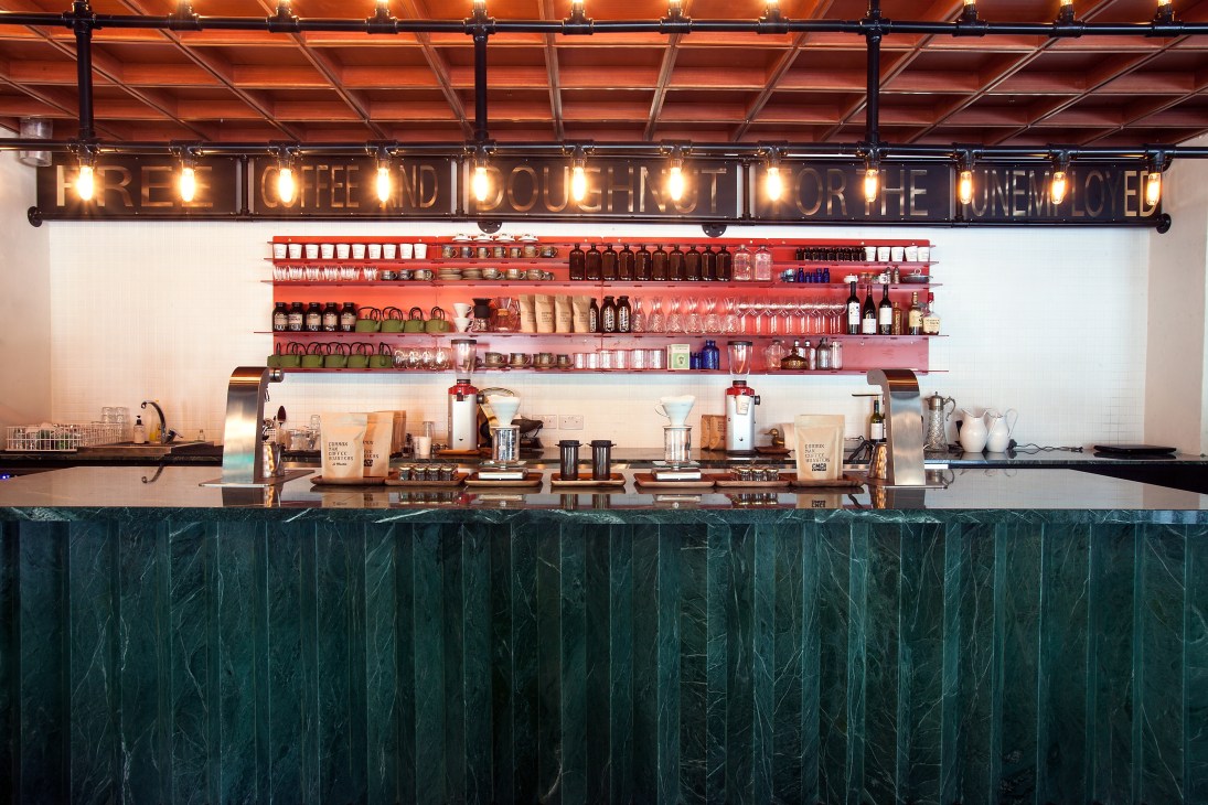 Common Man Coffee Roasters brew bar with espresso machines, shelving displaying coffee bags, and neon signage overhead.