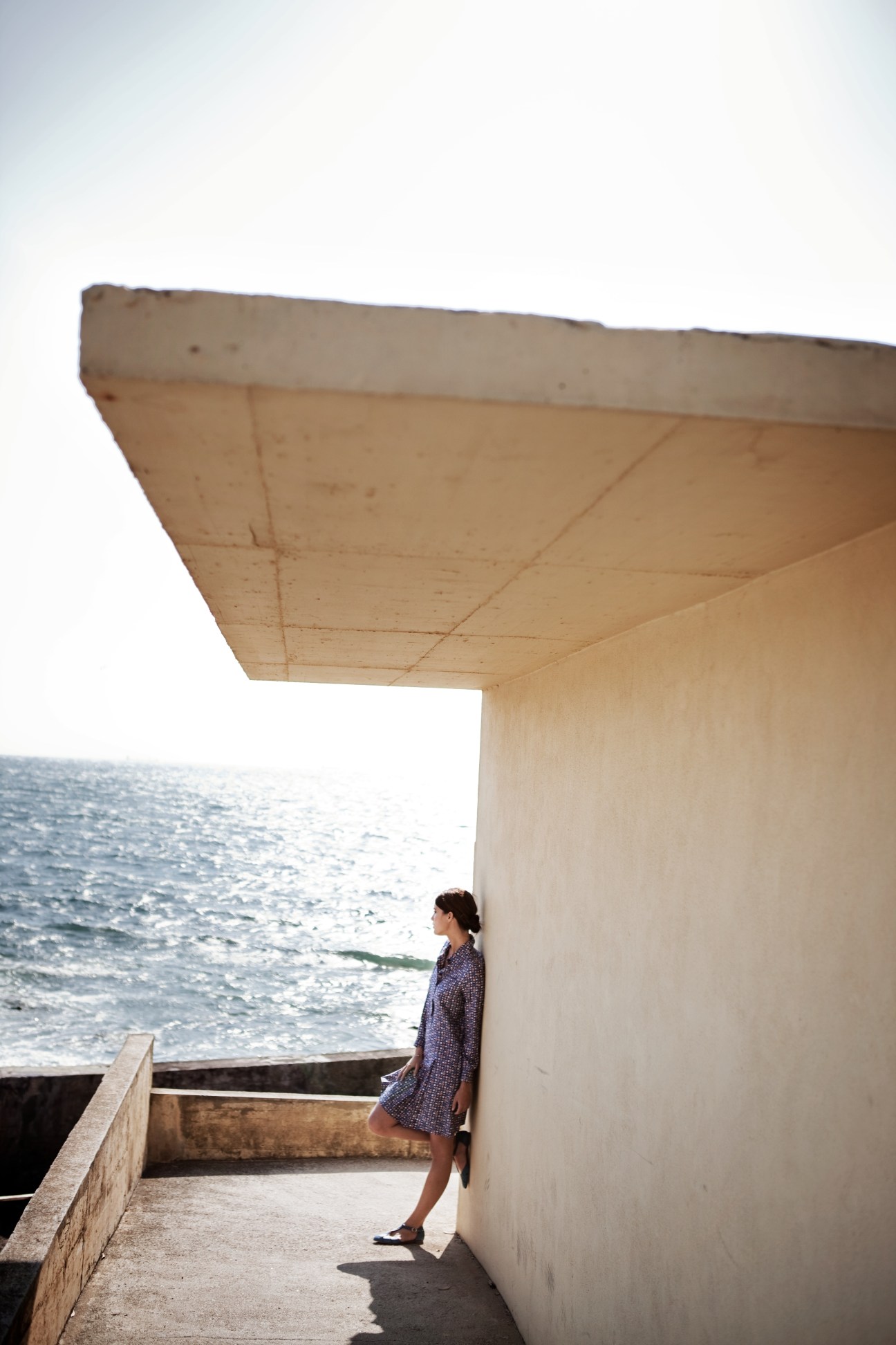 Woman wearing A.P.C. dress and Fratelli Rossetti shoes standing by concrete structure overlooking sea.