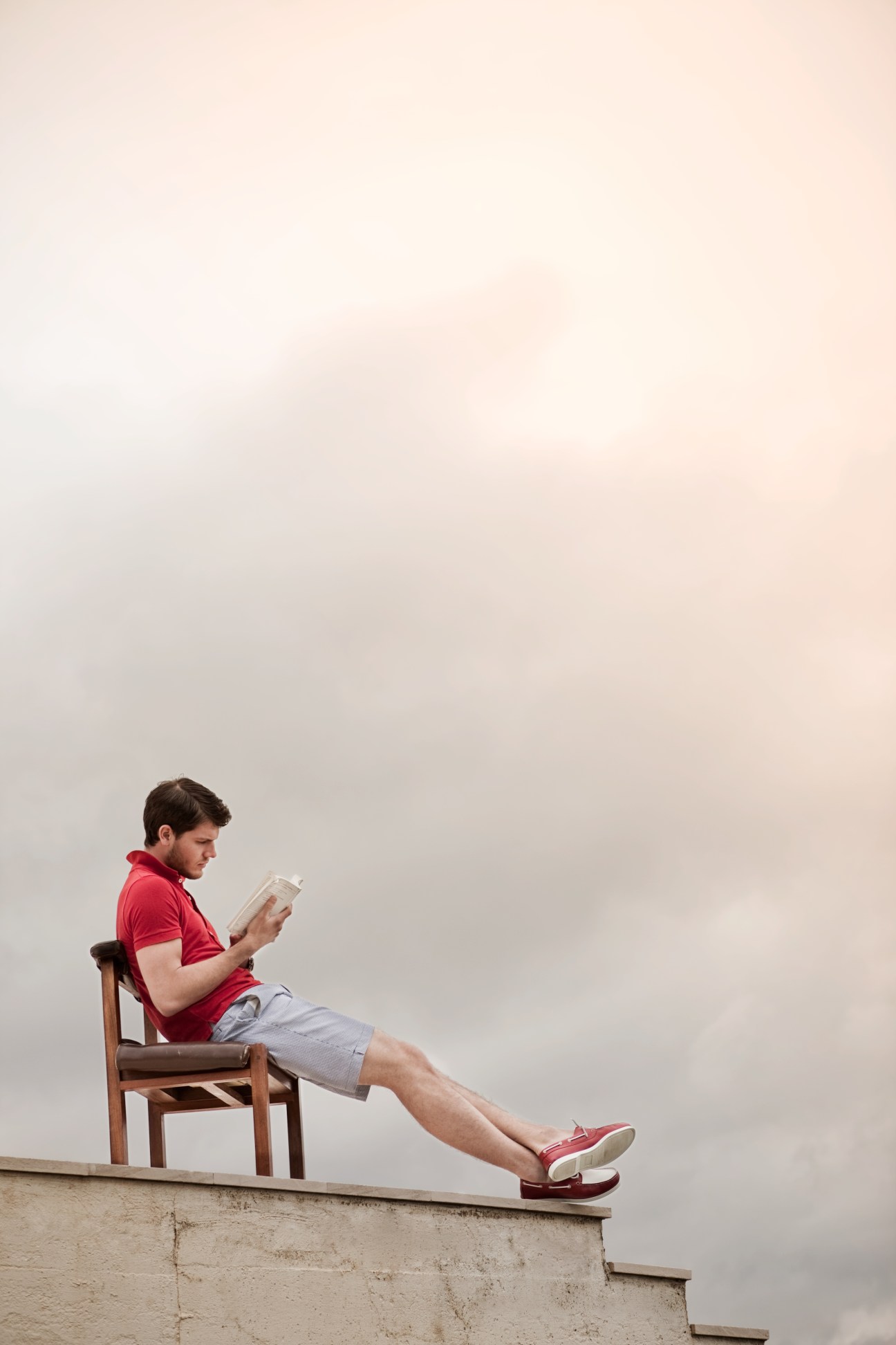 Man sitting on wooden chair wearing red polo shirt and shorts, reading on a rooftop overlooking the coast.
