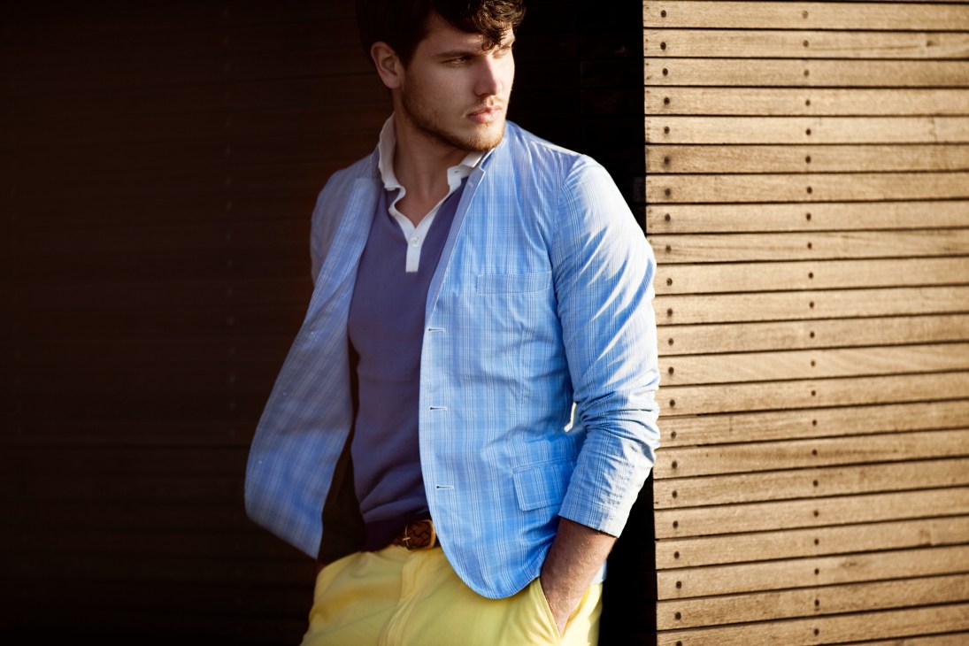 Man wearing light blue pinstriped jacket, navy polo shirt, and yellow shorts against wooden boardwalk backdrop.