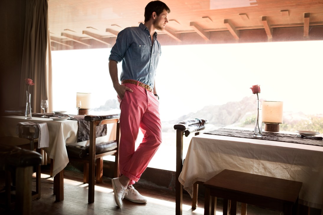 Man in denim shirt and burgundy trousers standing by a sunlit window with coastal views.