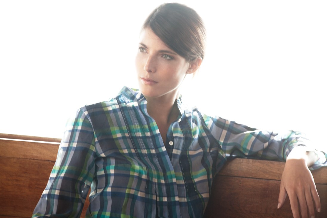 Woman wearing a blue and green plaid shirt by Glanshirt, seated against a wooden backdrop.